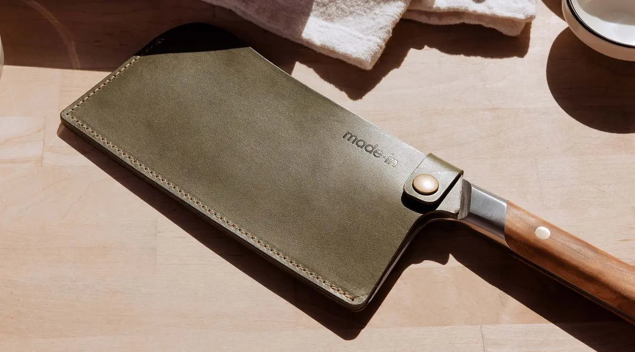 A chef's knife with a wooden handle and a protective leather sheath is resting on a wooden surface next to a towel and a bowl.
