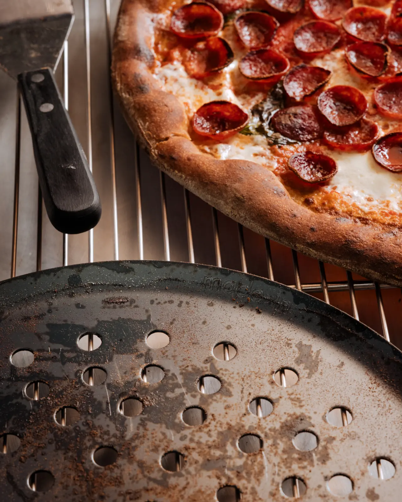 A freshly cooked pepperoni pizza rests on a cooling rack next to a pizza cutter and a worn pizza pan.