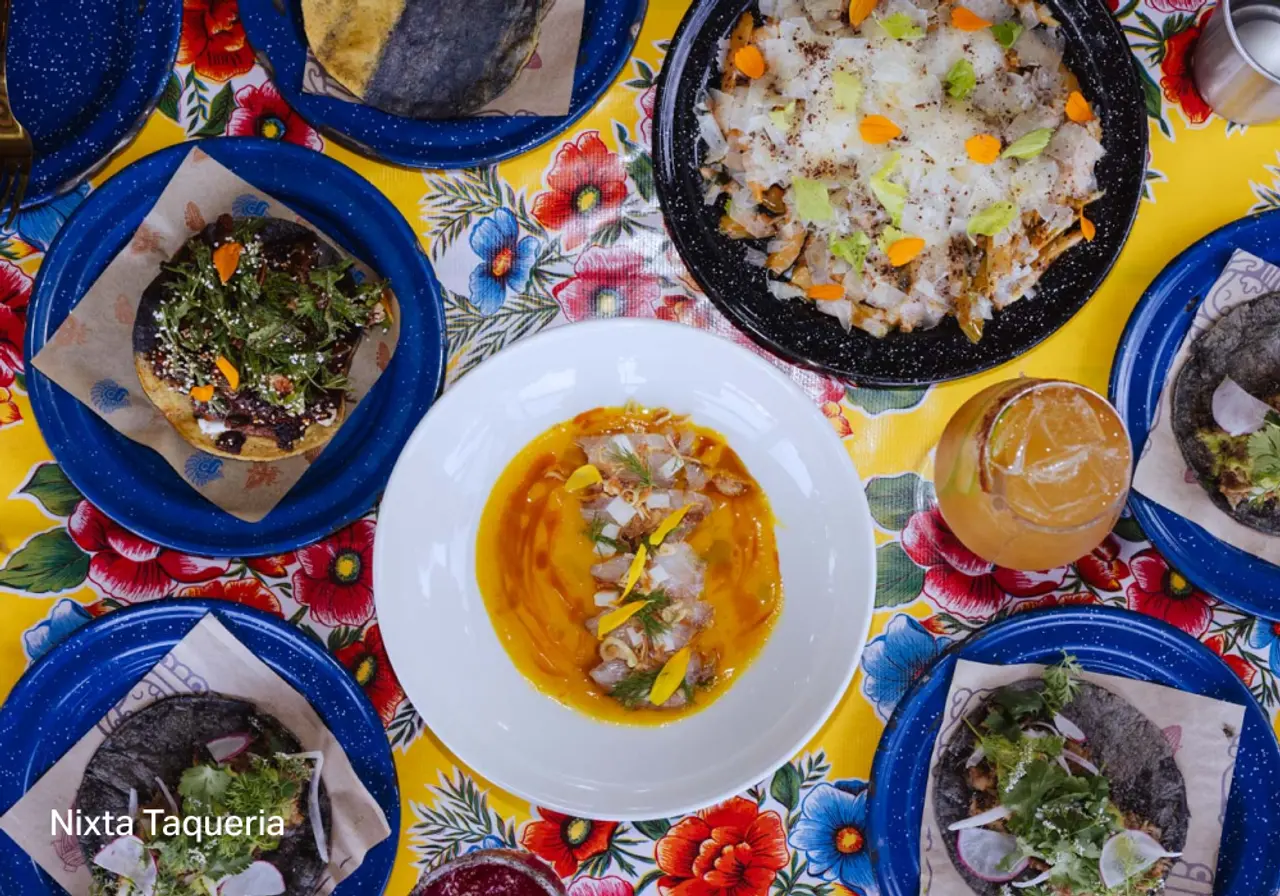 A spread of tacos, colorful dishes, and drinks sits on a floral tablecloth at Nixta Taqueria.