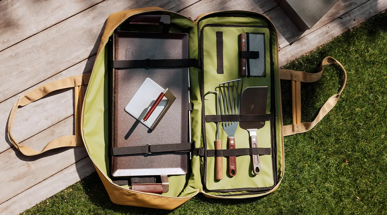 An open, organized grill set bag lies on the grass, containing various utensils like tongs, a spatula, and a fork, alongside a notebook and pens.