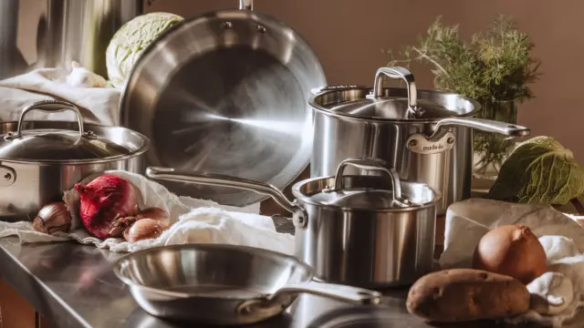 A collection of stainless steel cookware is arranged on a countertop alongside various vegetables and herbs.