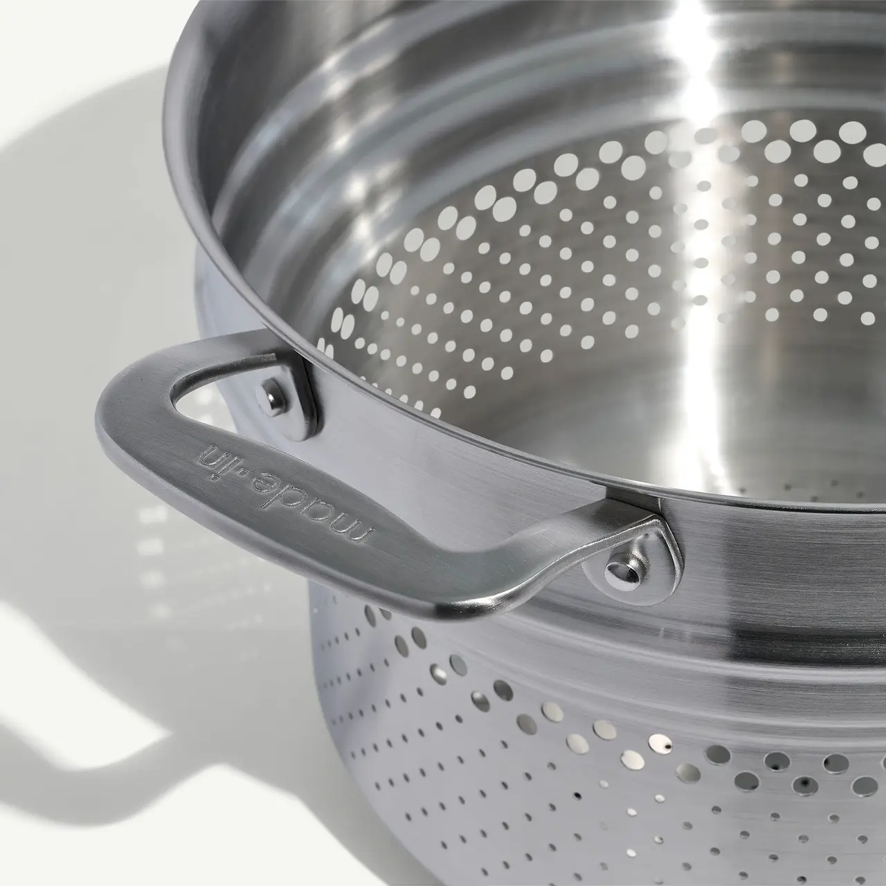 A close-up of a stainless steel colander showing its perforated surface and sturdy handle with a shadow cast on a plain background.