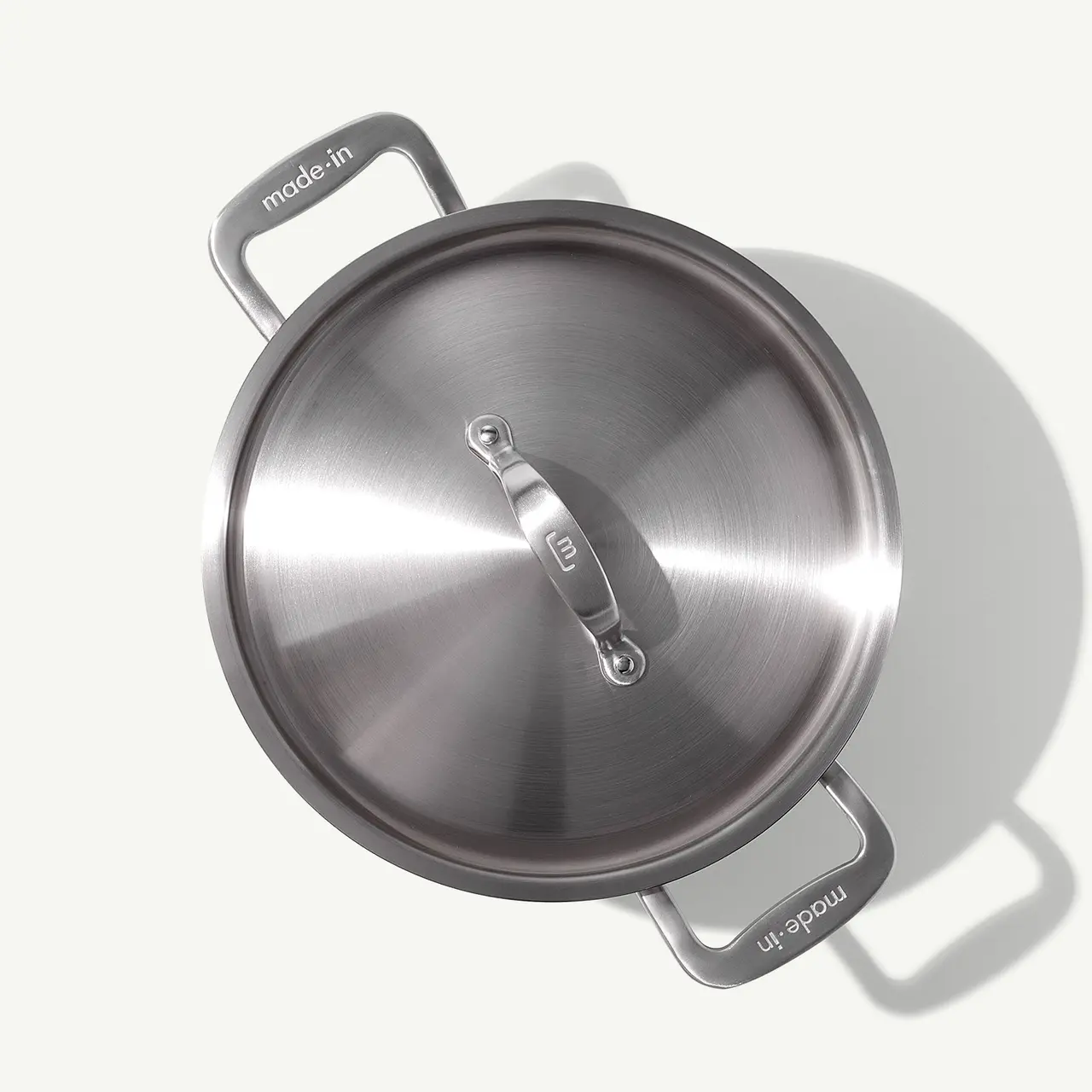 A stainless steel cooking pot with a lid is displayed from above, casting a soft shadow on a light surface.