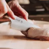 A hand holds a knife labeled "MADE IN JAPAN" while preparing raw chicken on a wooden cutting board.