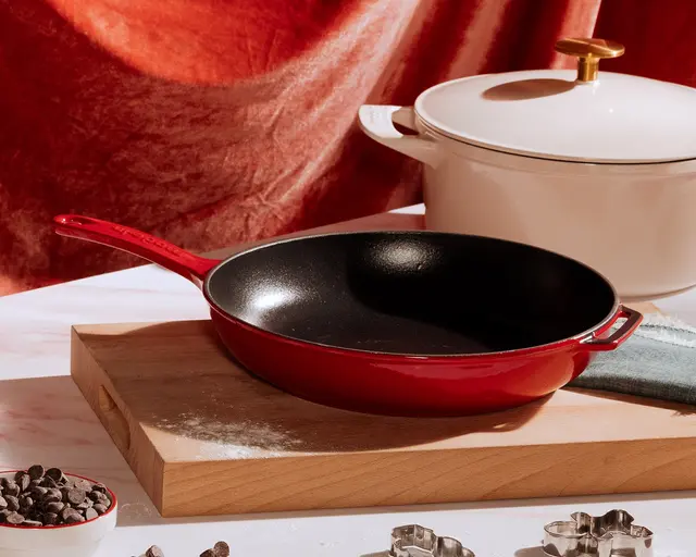 A red frying pan on a wooden cutting board with a white pot with a gold handle in the background, set against a warm-colored backdrop suggesting a cozy cooking atmosphere.
