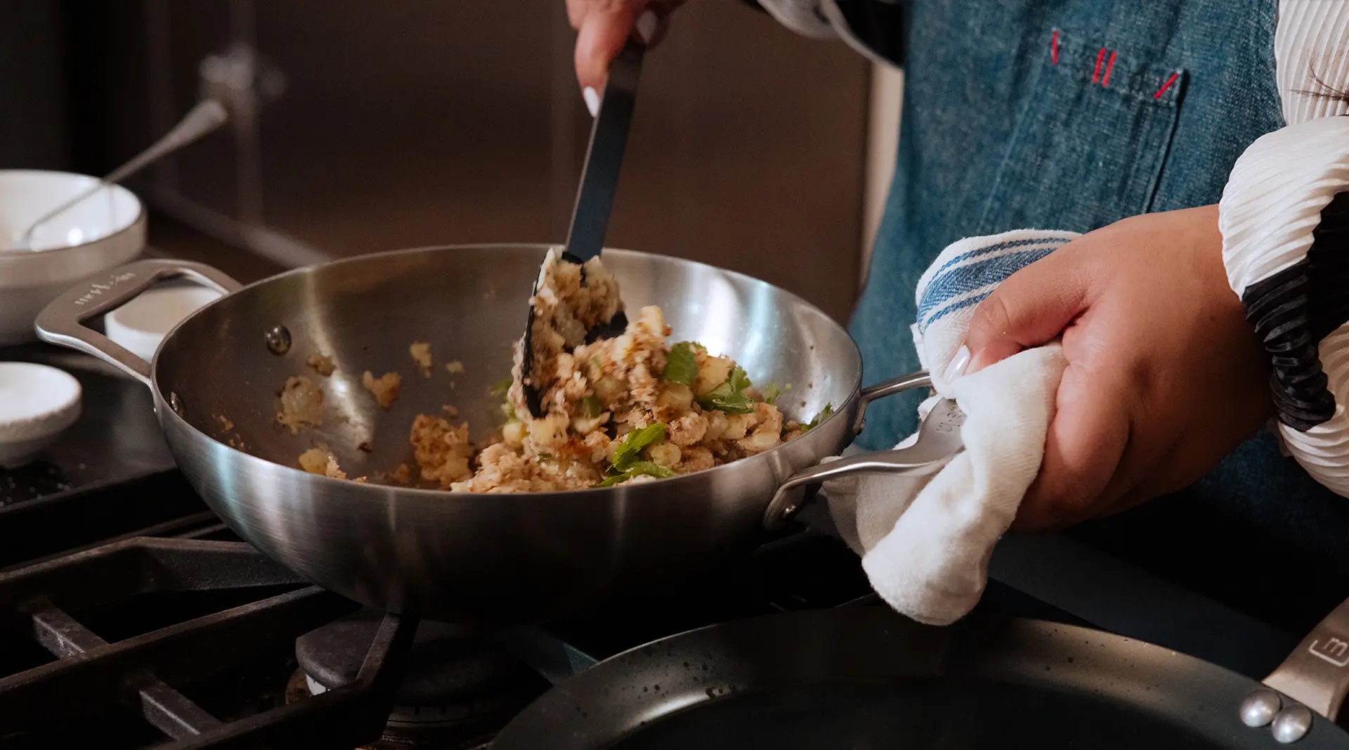 A person is cooking food in a frying pan on a stove, using a spatula and wearing an oven mitt.