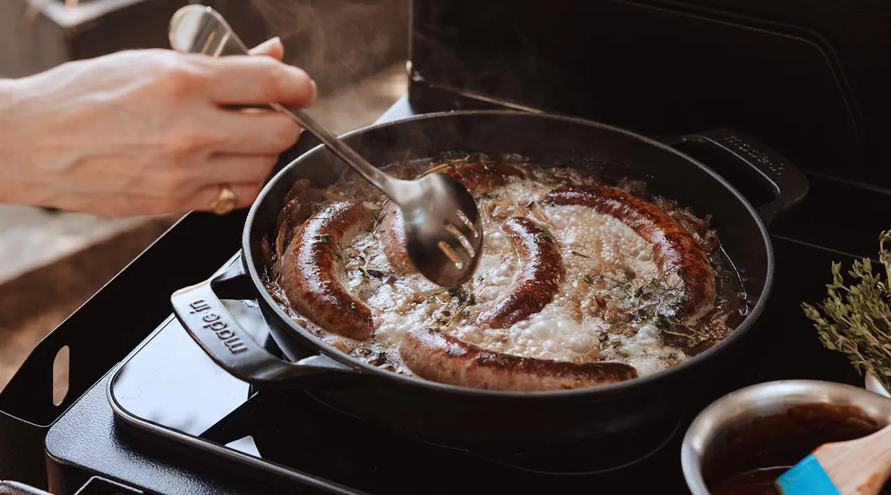 Sausages sizzle in a skillet on a stove as a person cooks, flipping them with a pair of tongs.