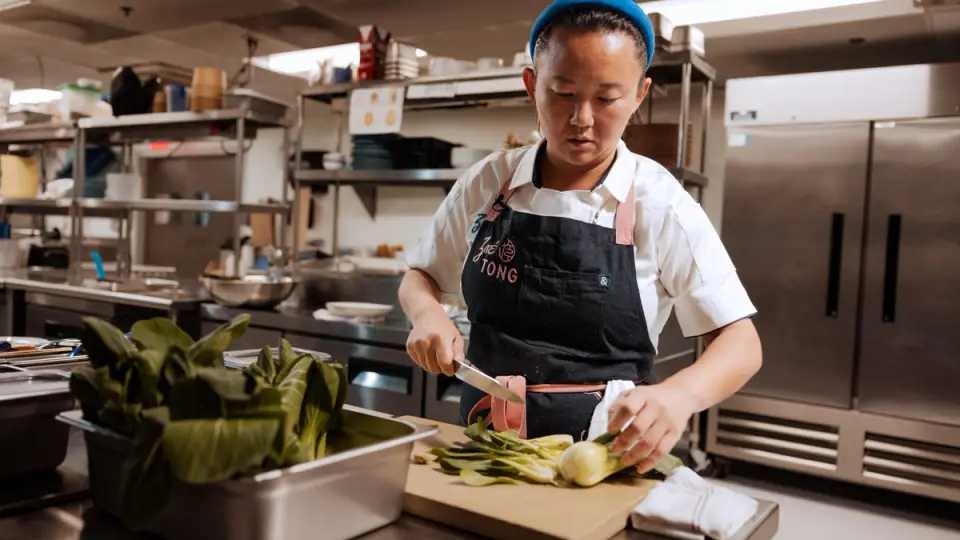 A focused chef wearing an apron trims vegetables on a cutting board in a professional kitchen.