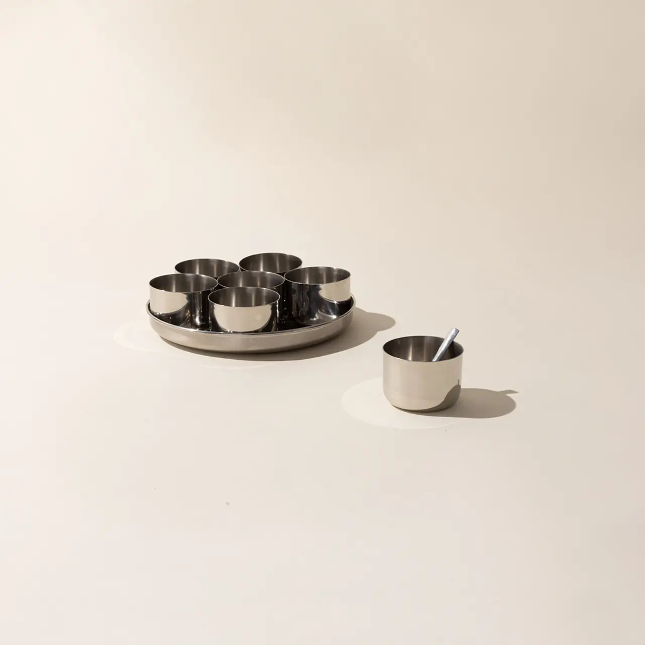 A set of stainless steel cups are neatly placed on a tray with one cup sitting isolated from the others on a plain background.