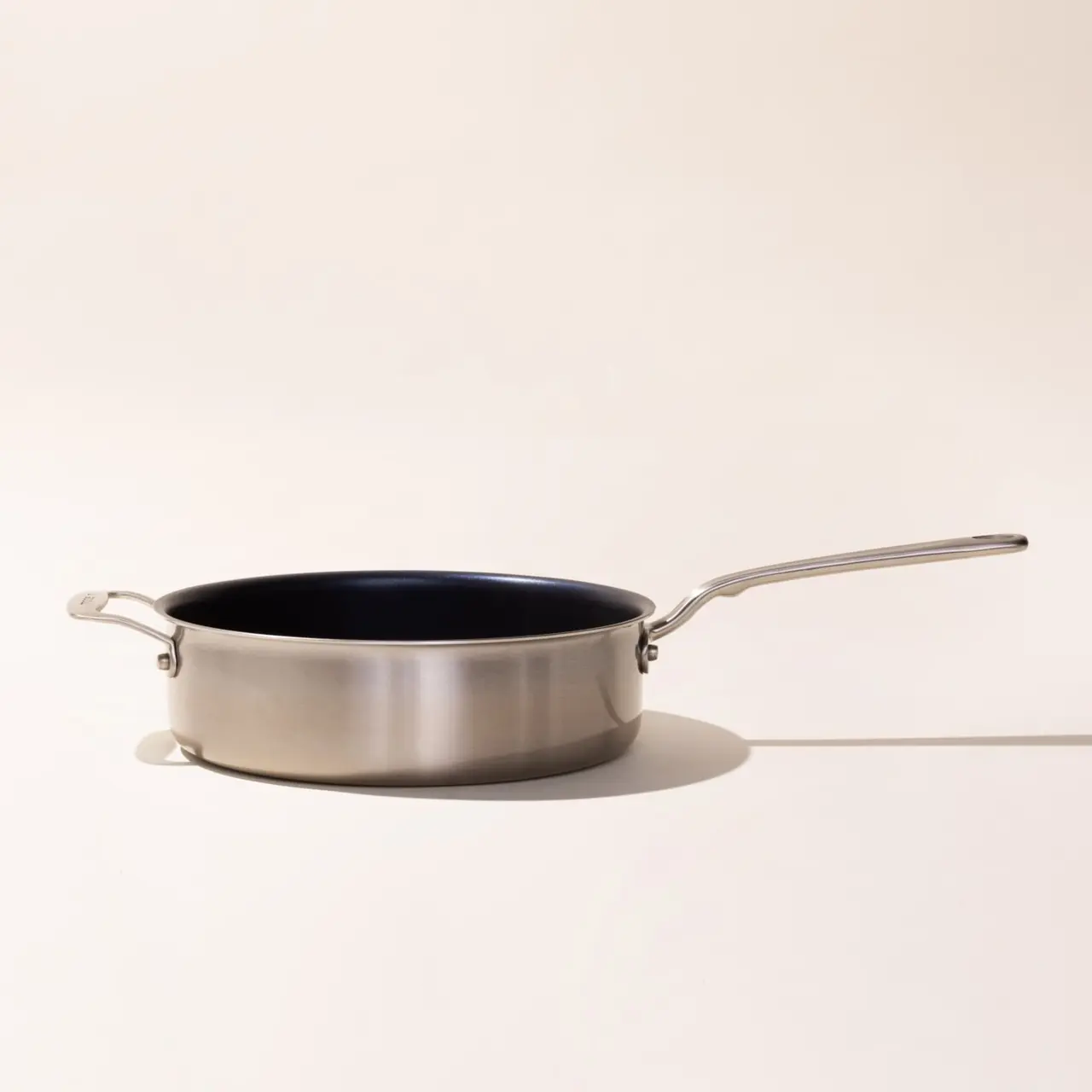 A stainless steel frying pan with a long handle is positioned on a plain surface with a neutral background.