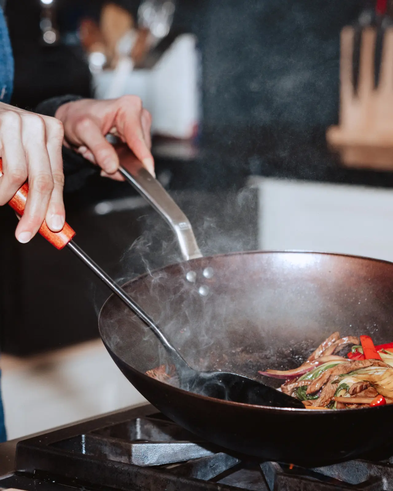 A person is using a spatula to stir-fry vegetables in a hot wok while steam rises from the pan.