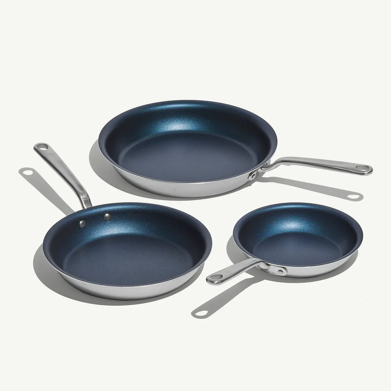 Three different-sized frying pans with a metallic exterior and blue interior sit on a light surface.