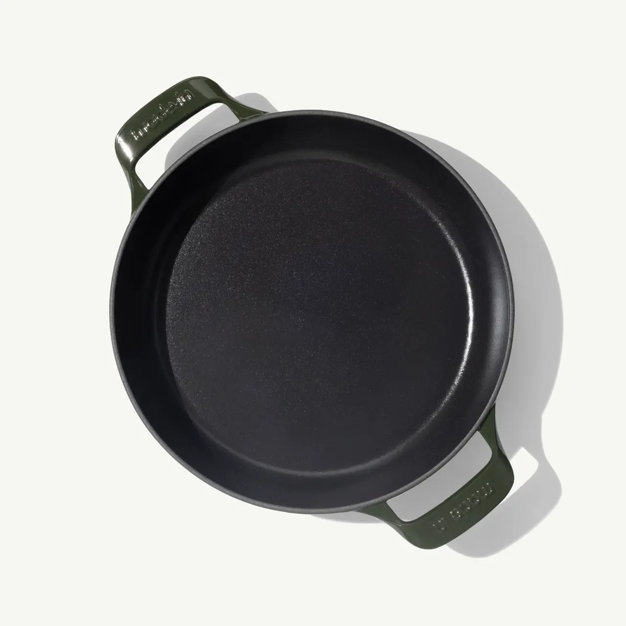 A round, black cast iron skillet with green handles is shown from a top-down perspective.