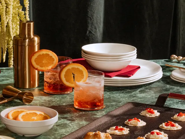 A table set with cocktails, a gold shaker, stacked plates, a bowl of orange slices, and canapés, creating an elegant dining atmosphere.