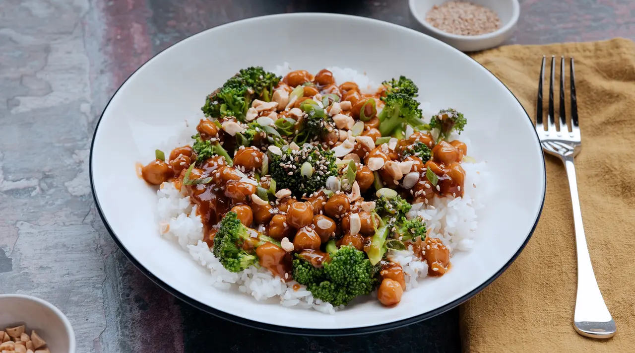 A plate of rice topped with broccoli, chickpeas, and sesame seeds drizzled with sauce is placed on a table next to a fork and a small bowl of seeds.