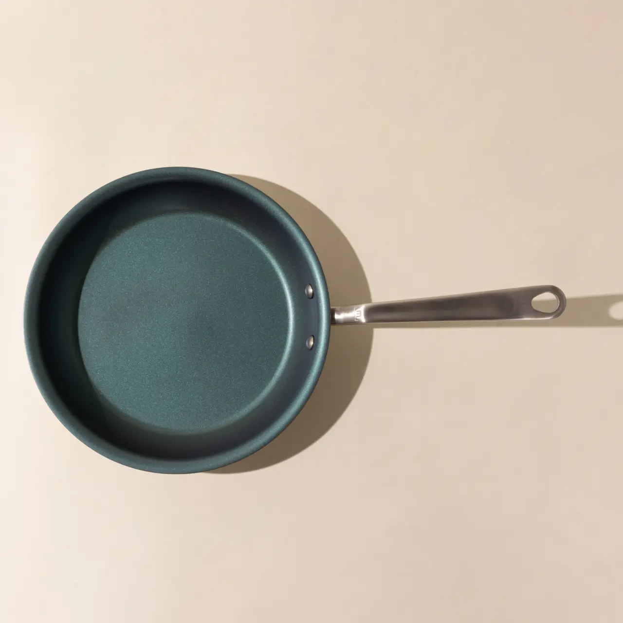A non-stick frying pan with a stainless steel handle is shown from above, casting a slight shadow on a beige surface.