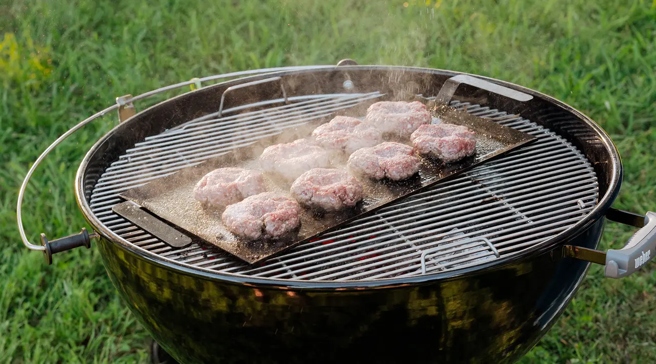 A lineup of raw burger patties grills on a salt block atop a round barbecue on a grassy background.