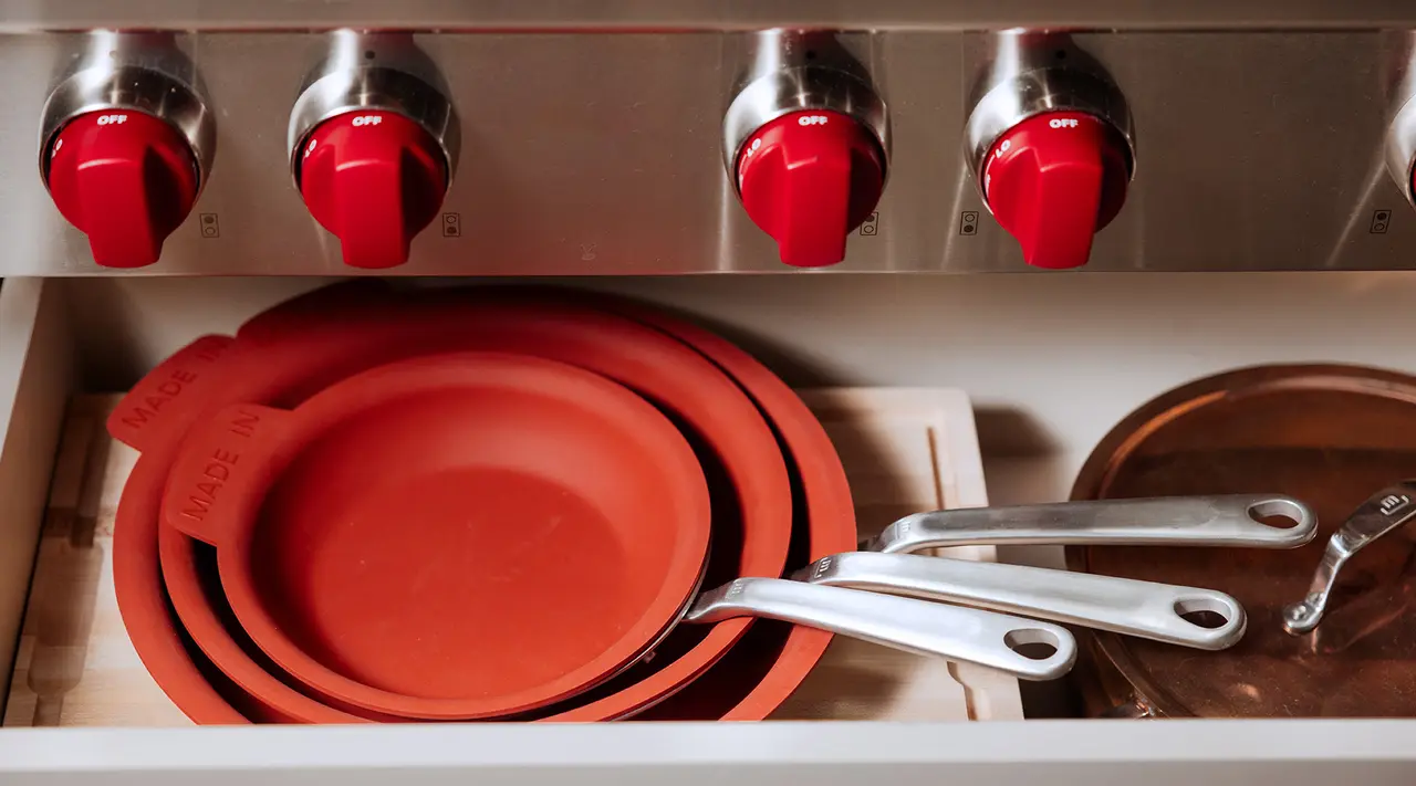 A set of red silicone lids is neatly stored next to stainless steel pans in a kitchen drawer, with stove knobs visible above.