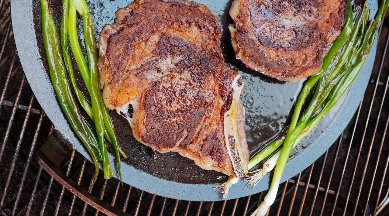 Two juicy steaks with grill marks alongside asparagus spears are cooking on a cast-iron pan over a grill.