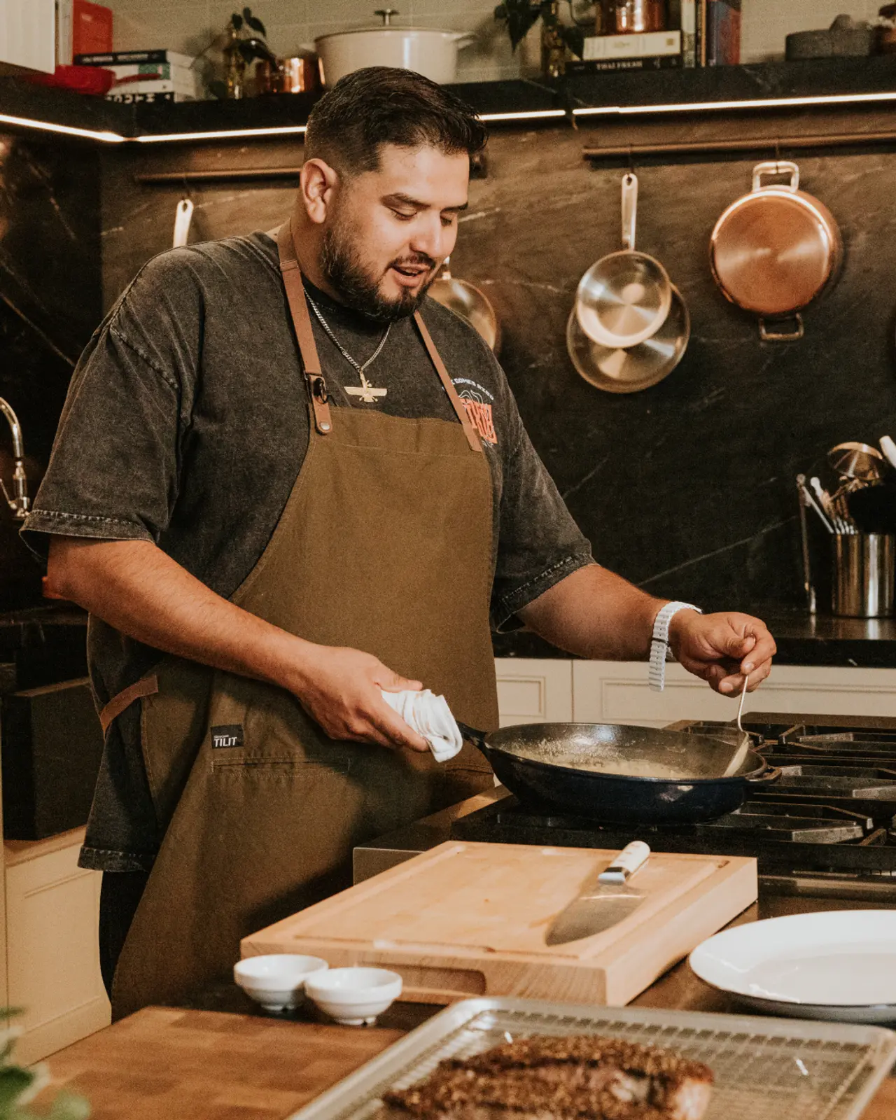 A focused chef in an apron is sautéing food in a pan amidst a kitchen setting adorned with copper pots.