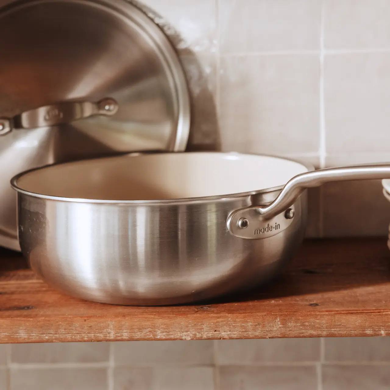 A stainless steel pot with a beige interior sits on a wooden shelf, accompanied by its matching lid in the background.