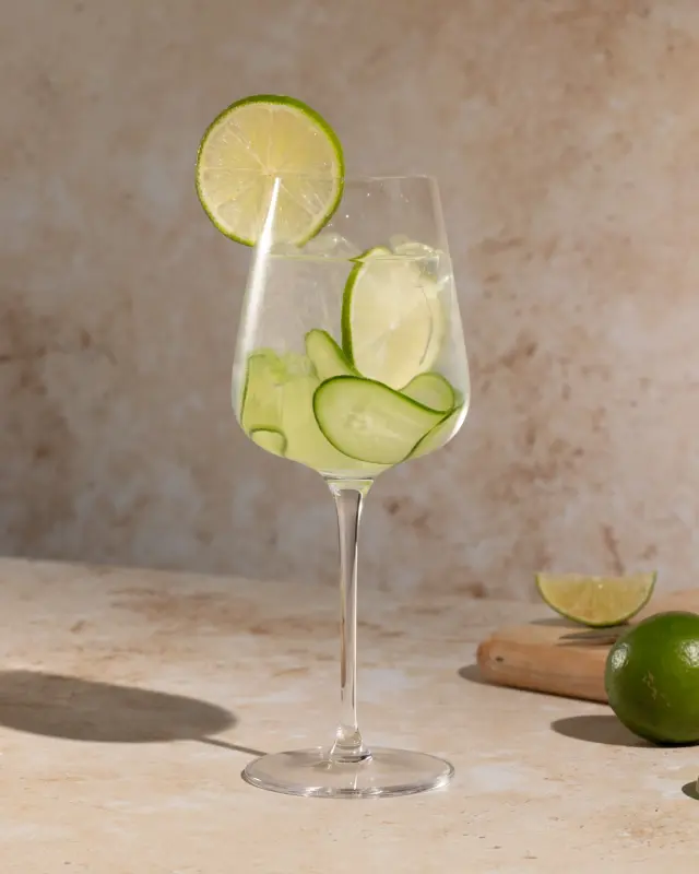 A refreshing glass of beverage with ice cubes, cucumber slices, and a lime wheel, situated on a sunlit surface with lime pieces nearby.