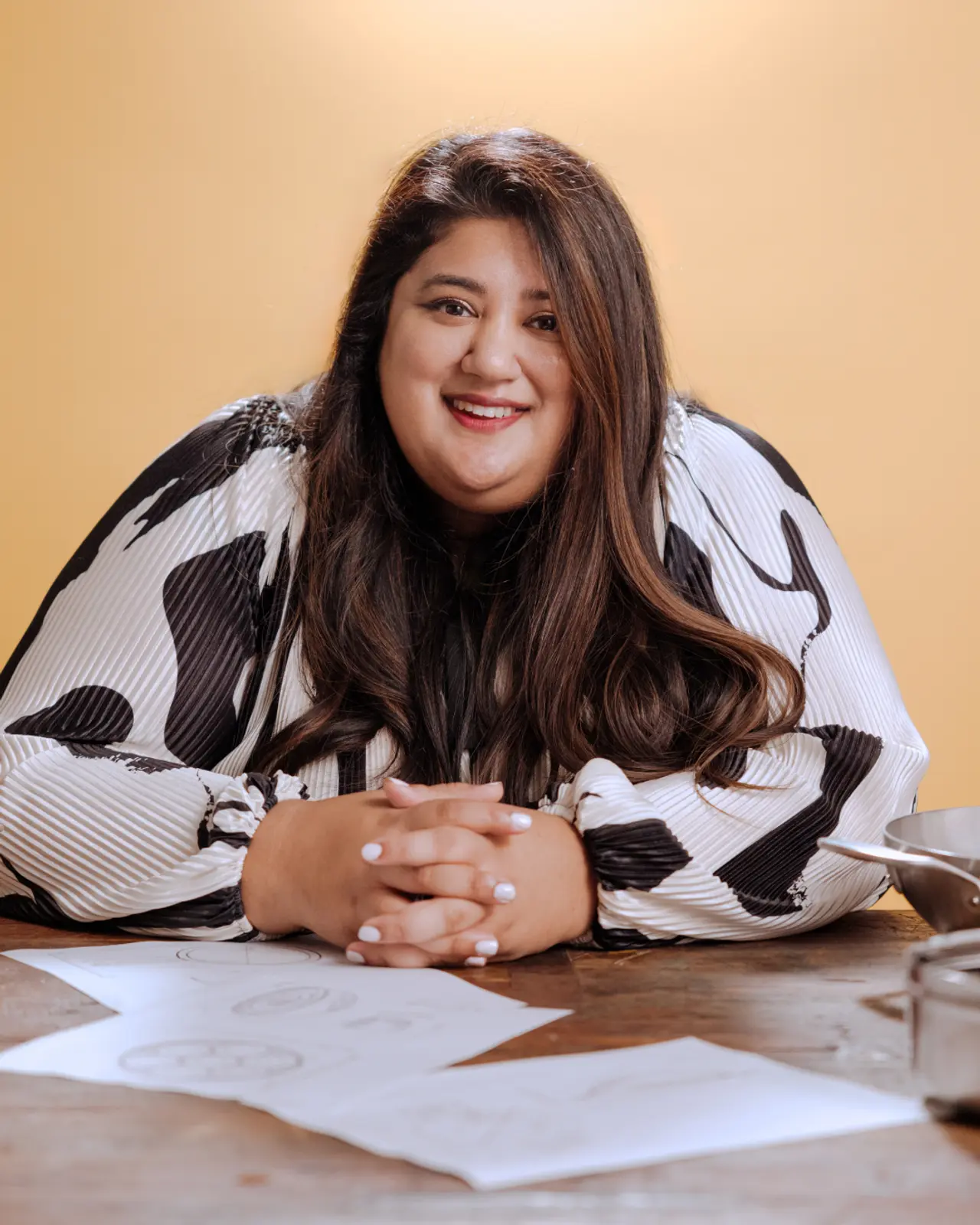A smiling person with long hair is sitting at a table with papers in front of them, wearing a black and white patterned top.