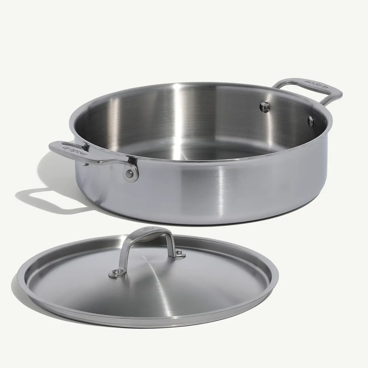 A stainless steel sauté pan with its lid placed to the side, showing reflective surfaces and sturdy handles.