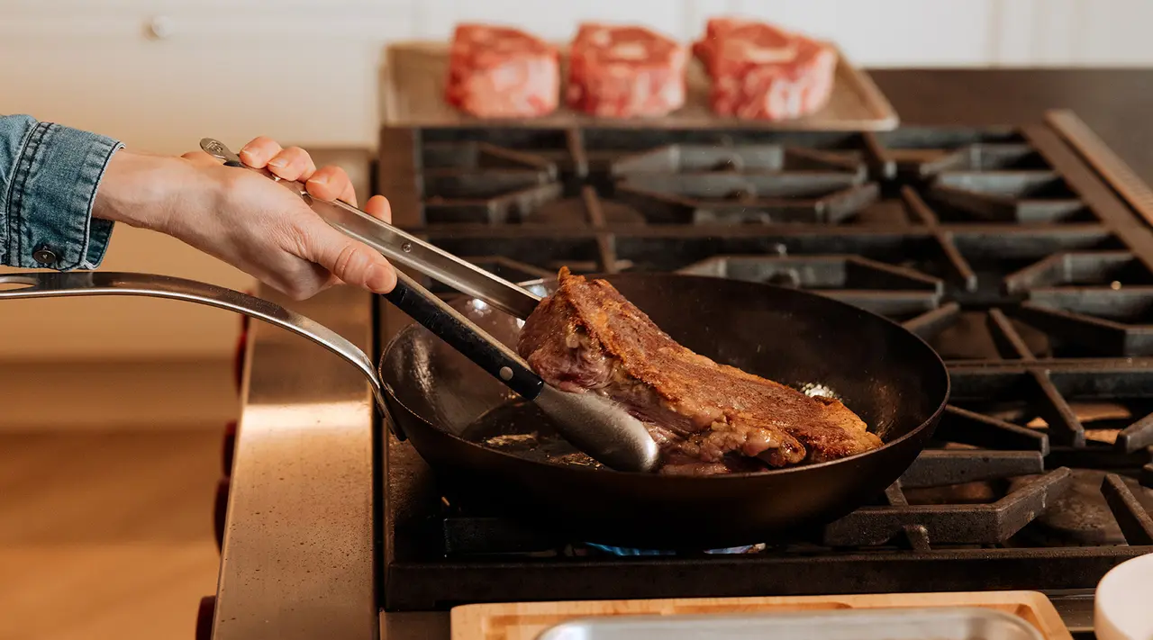 A person is searing a large steak in a skillet on a stove, with uncooked steaks visible in the background.