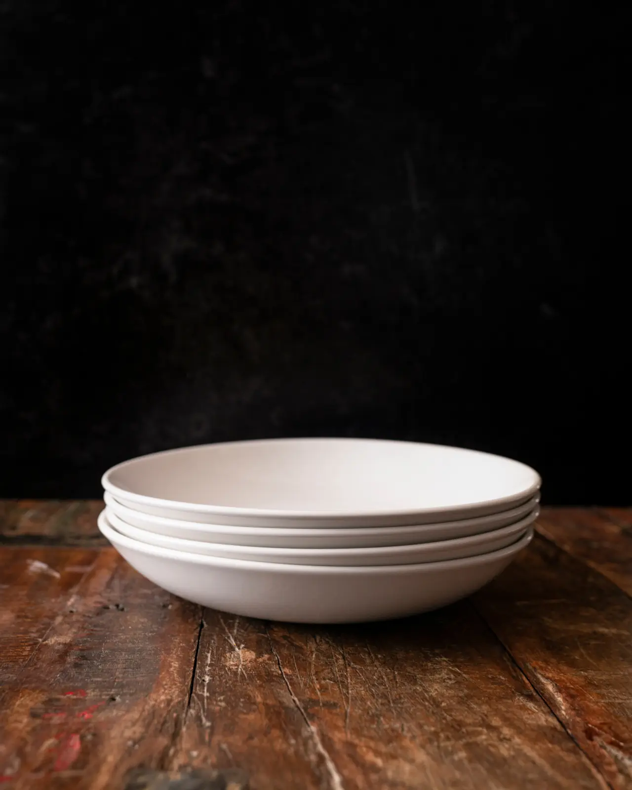 A stack of white plates is positioned on a worn wooden table against a dark background.