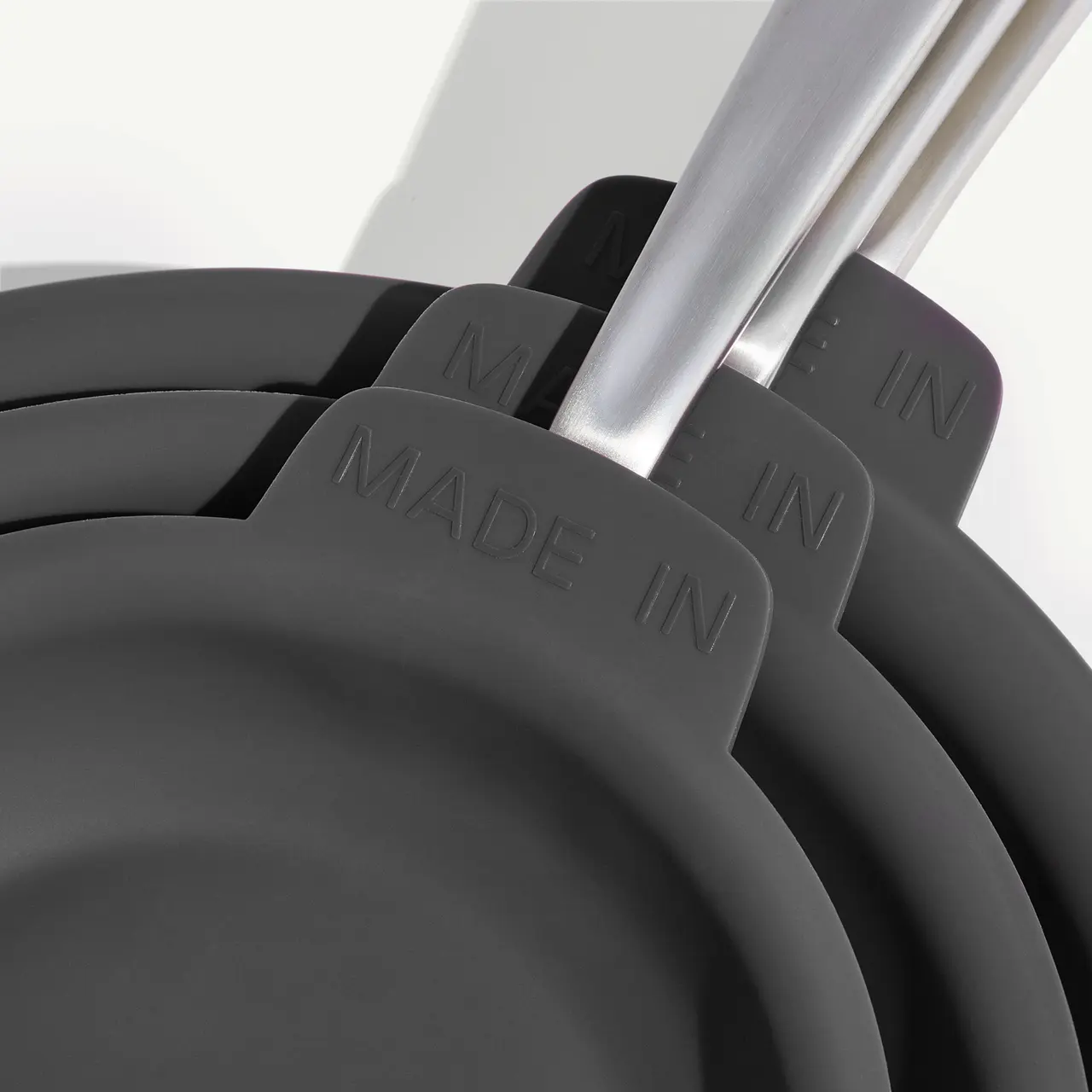 A close-up of a stack of dark non-stick pans with "MADE IN" imprinted on their handles, suggesting a focus on the manufacturing origin.