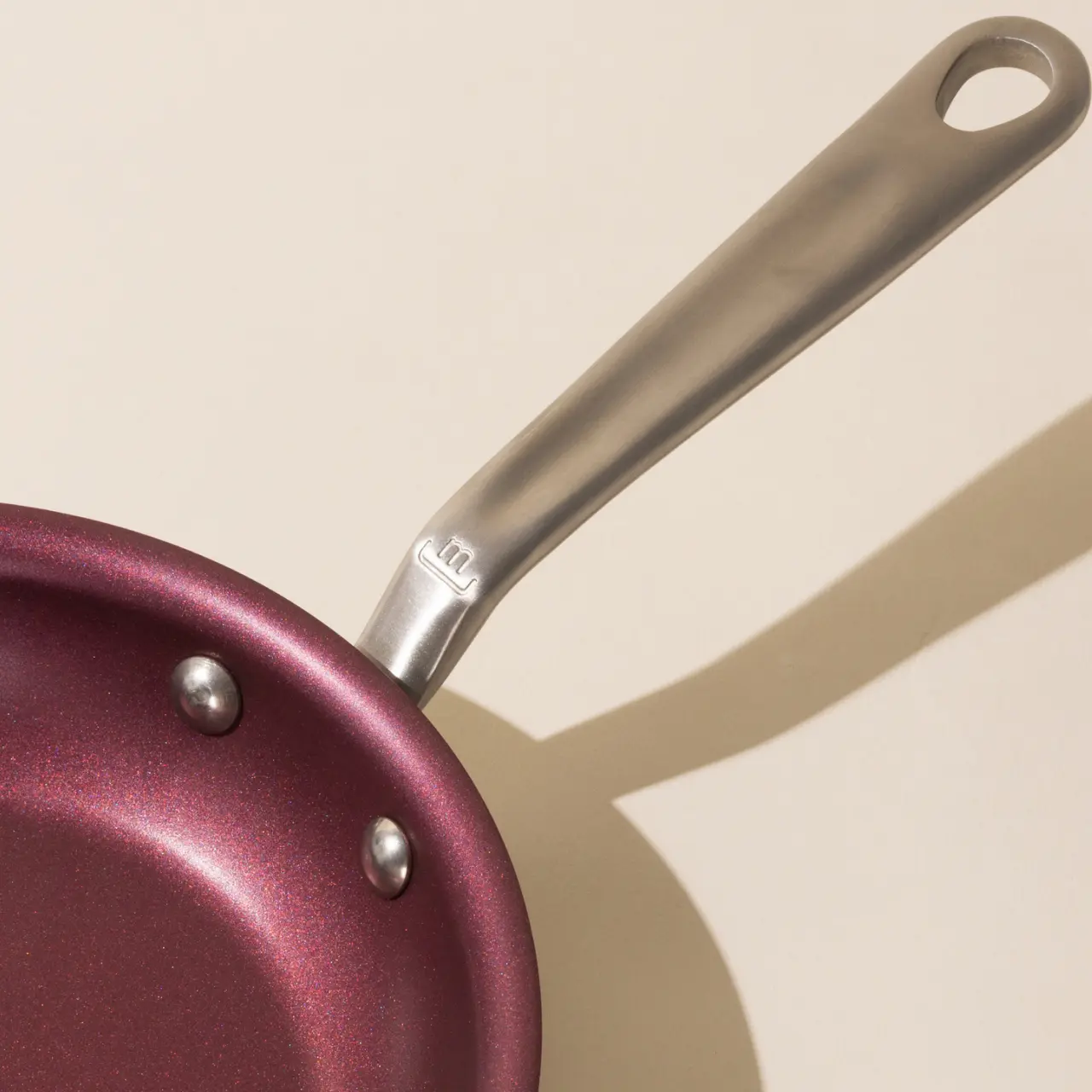 A burgundy non-stick frying pan with a silver handle cast a shadow on a light surface.