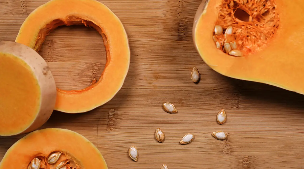 Sliced butternut squash and seeds are scattered on a wooden surface.