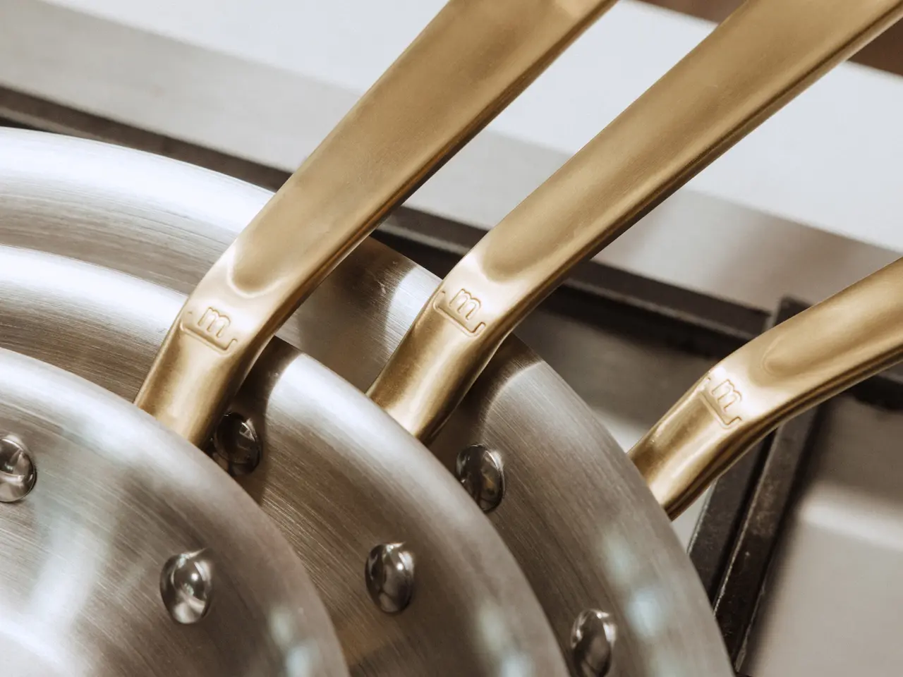 Three stainless steel frying pans with gold-toned handles are stacked on a stove.