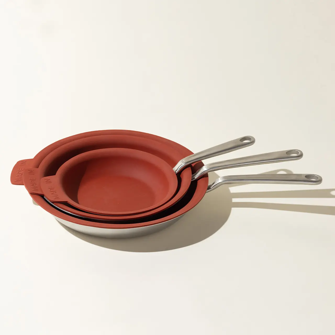 A stack of three red frying pans with silver handles casts a long shadow on a light surface.