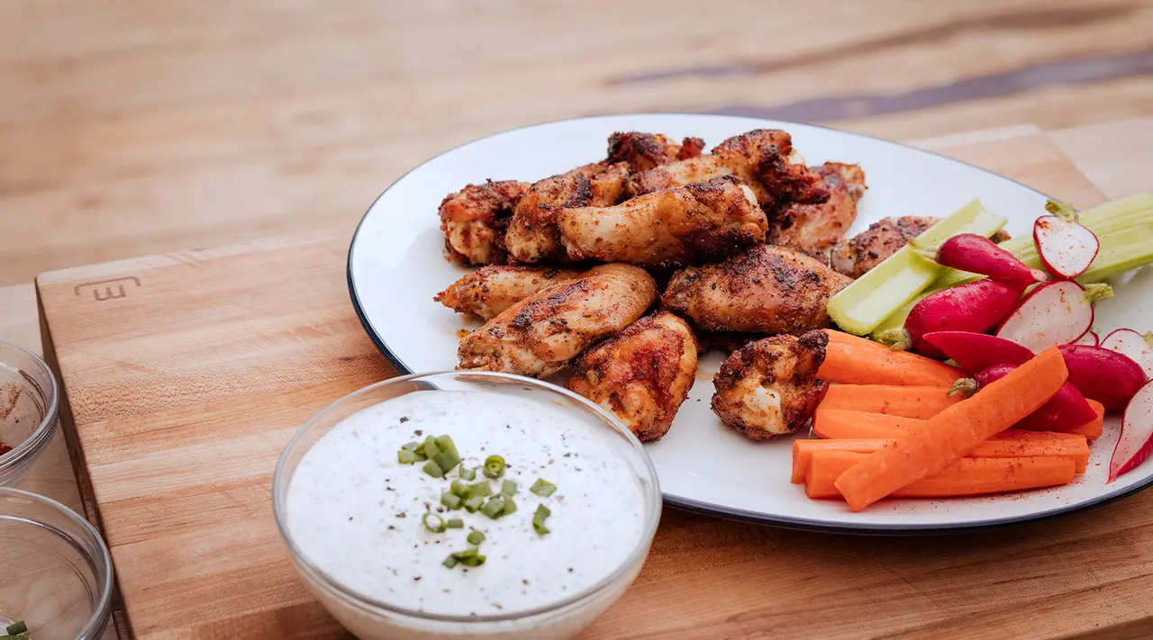 Grilled chicken wings on a plate with a side of fresh carrot sticks, celery, and radishes, accompanied by a bowl of dip.