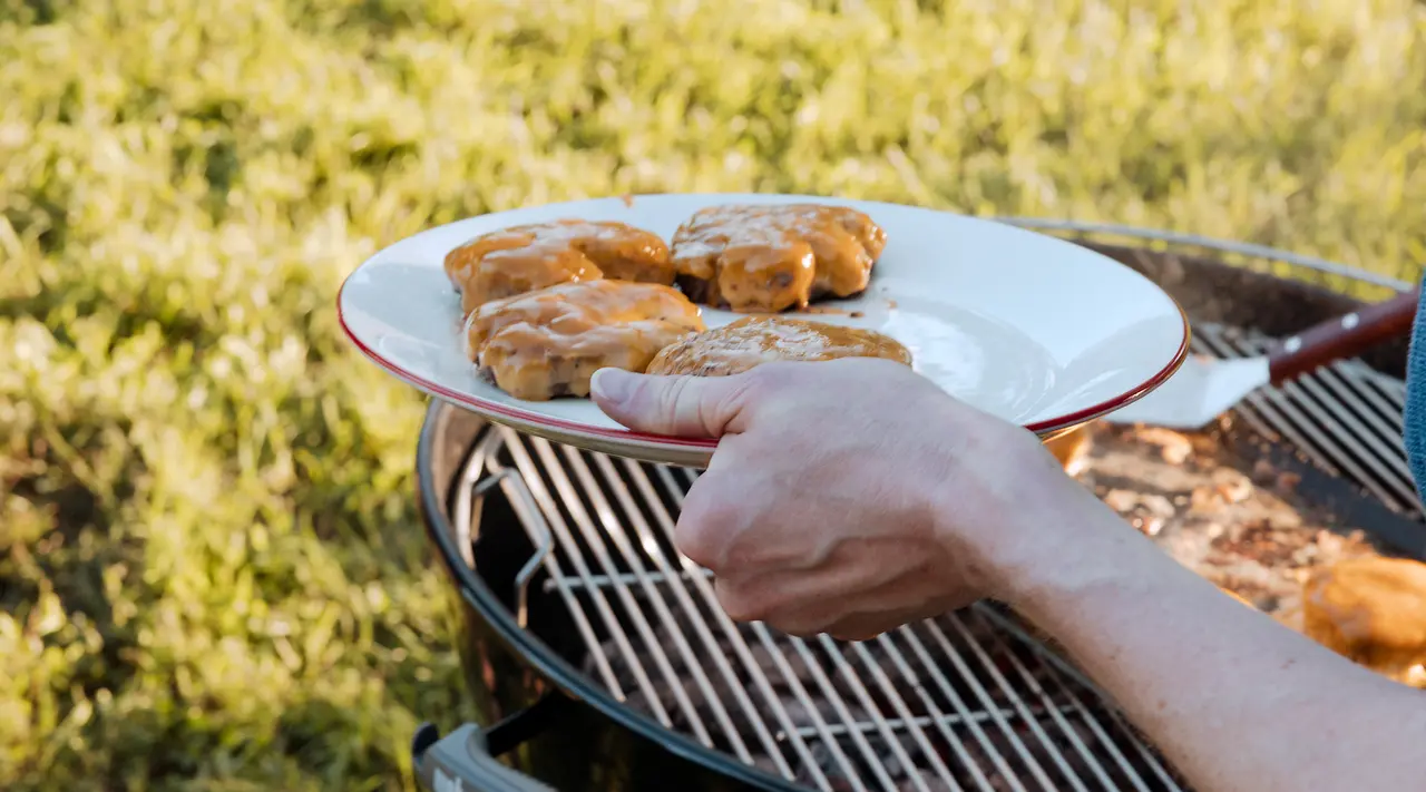 A person's hand is holding a plate with grilled chicken breasts over a barbecue grill.