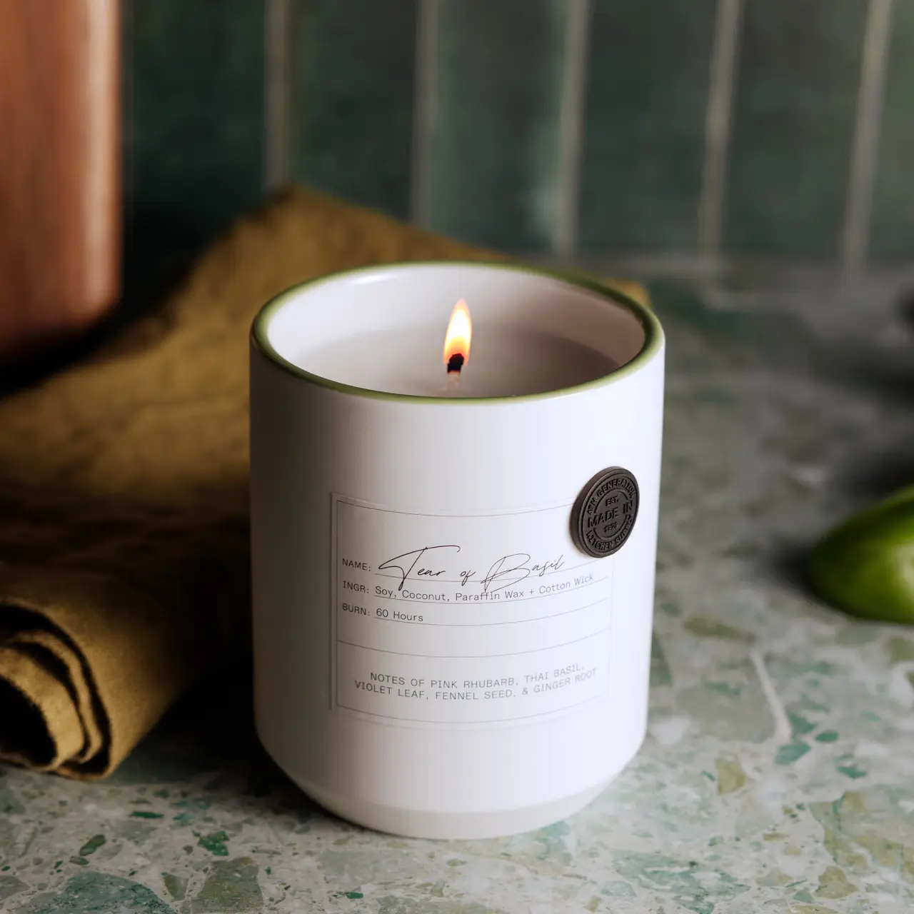A lit scented candle with a label is placed on a countertop near a mustard-colored cloth and a hint of green limes in the background.