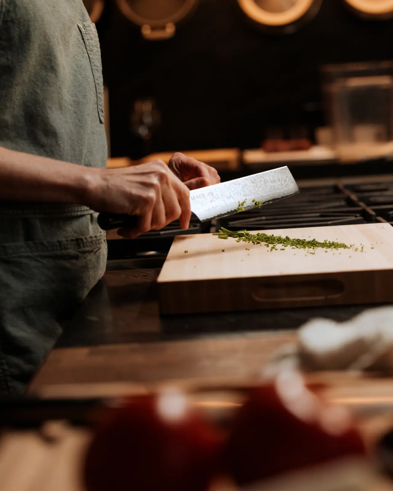 A person in an apron is meticulously chopping herbs on a wooden cutting board in a kitchen setting.