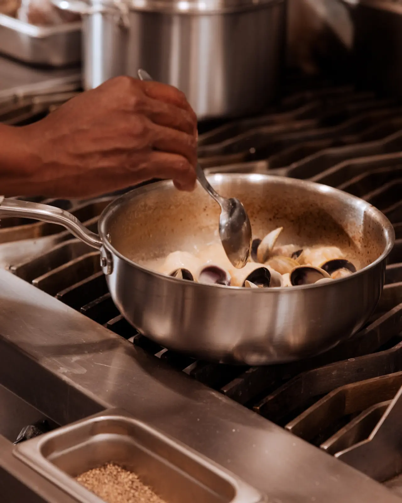 A hand stirs a pot of clams cooking on a stovetop in a modern kitchen.