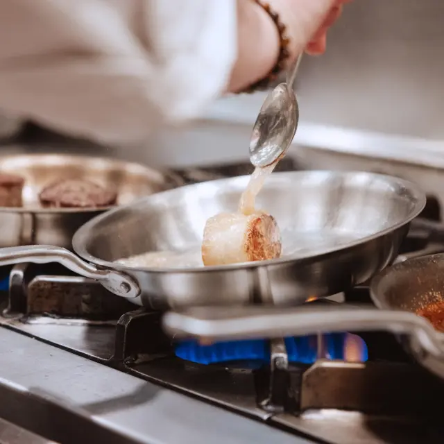A hand pours a sauce from a spoon into a simmering pan on a stovetop, with other pans visible in the background.