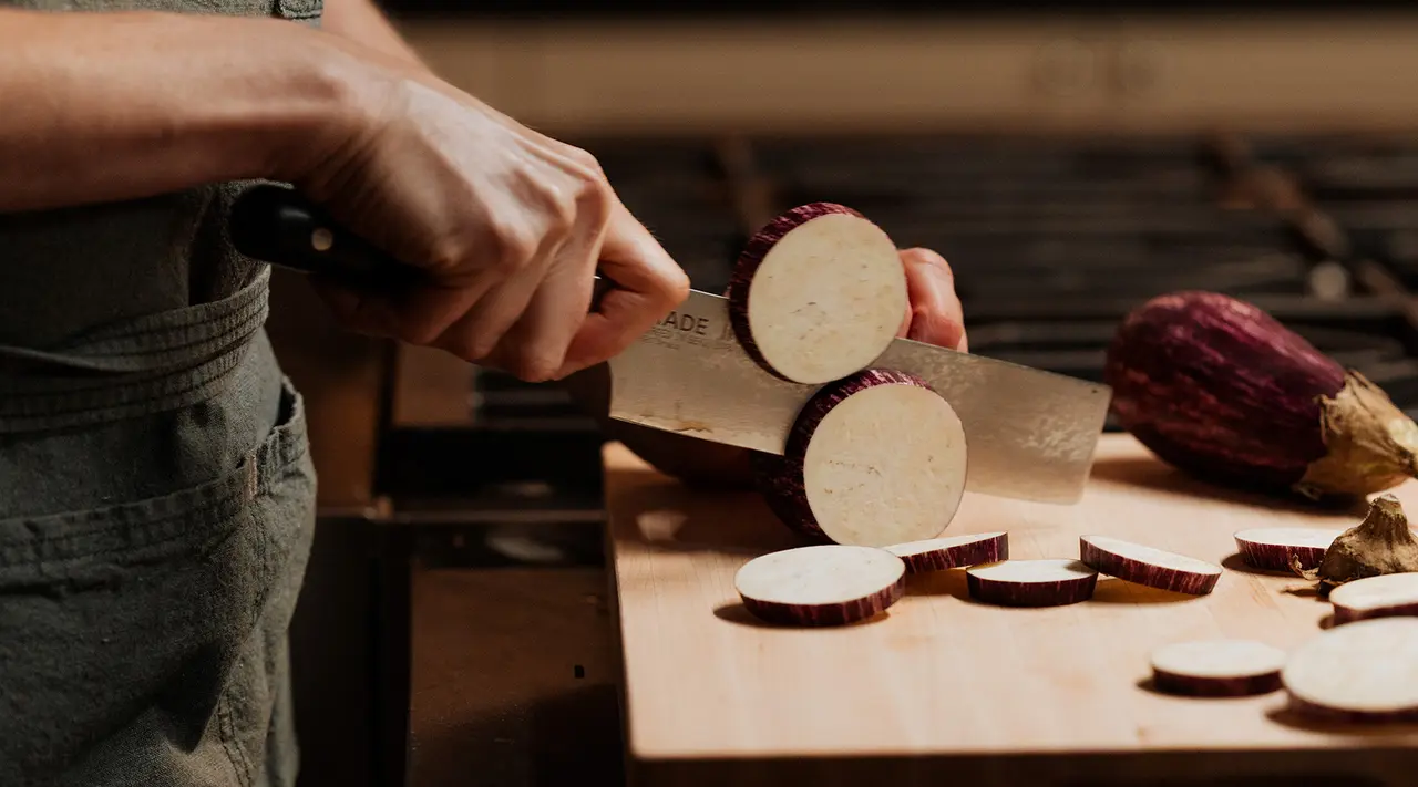 A person is slicing an eggplant on a wooden cutting board.