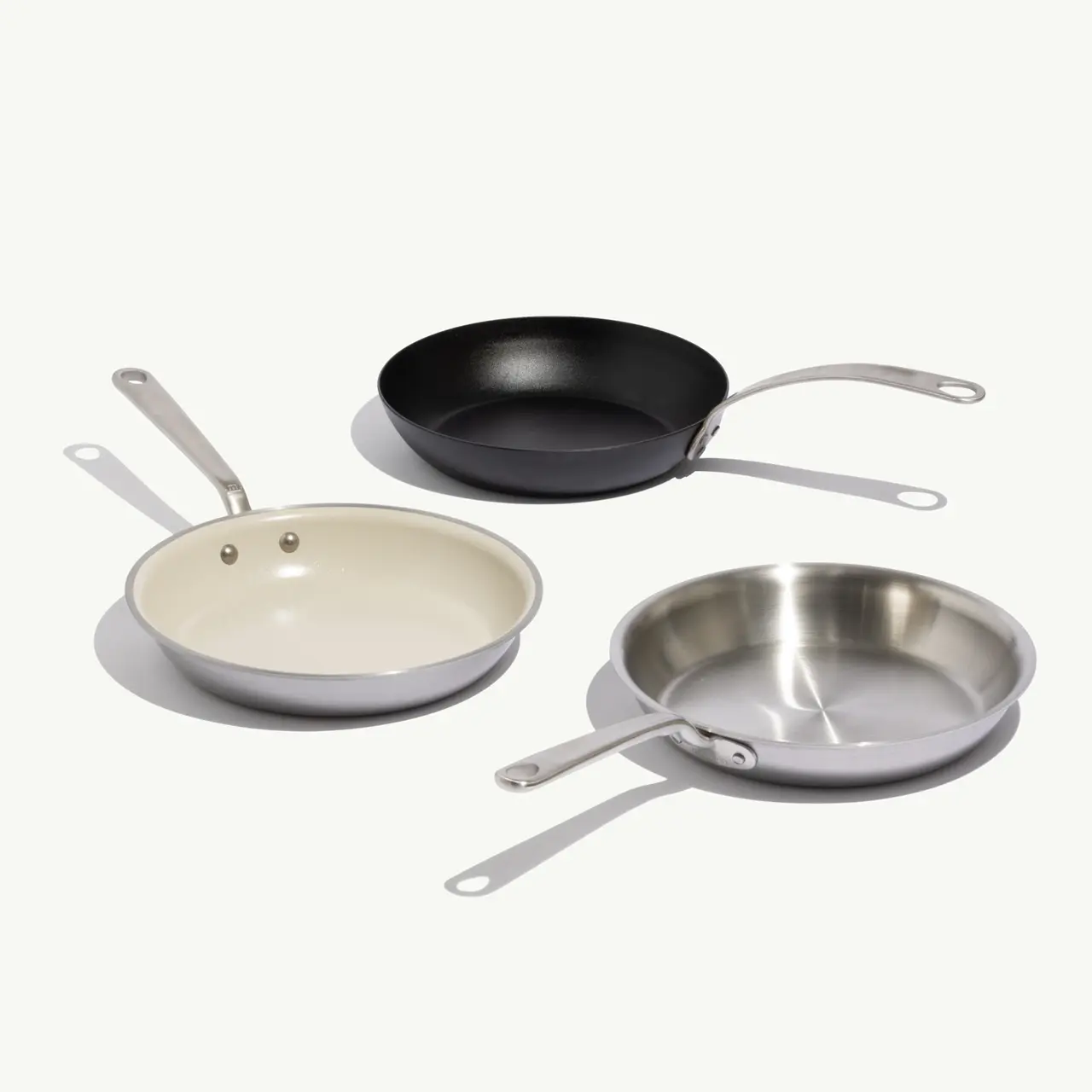 Three frying pans in different materials sit on a light background: a black non-stick pan, a white ceramic-coated pan, and a stainless steel pan.