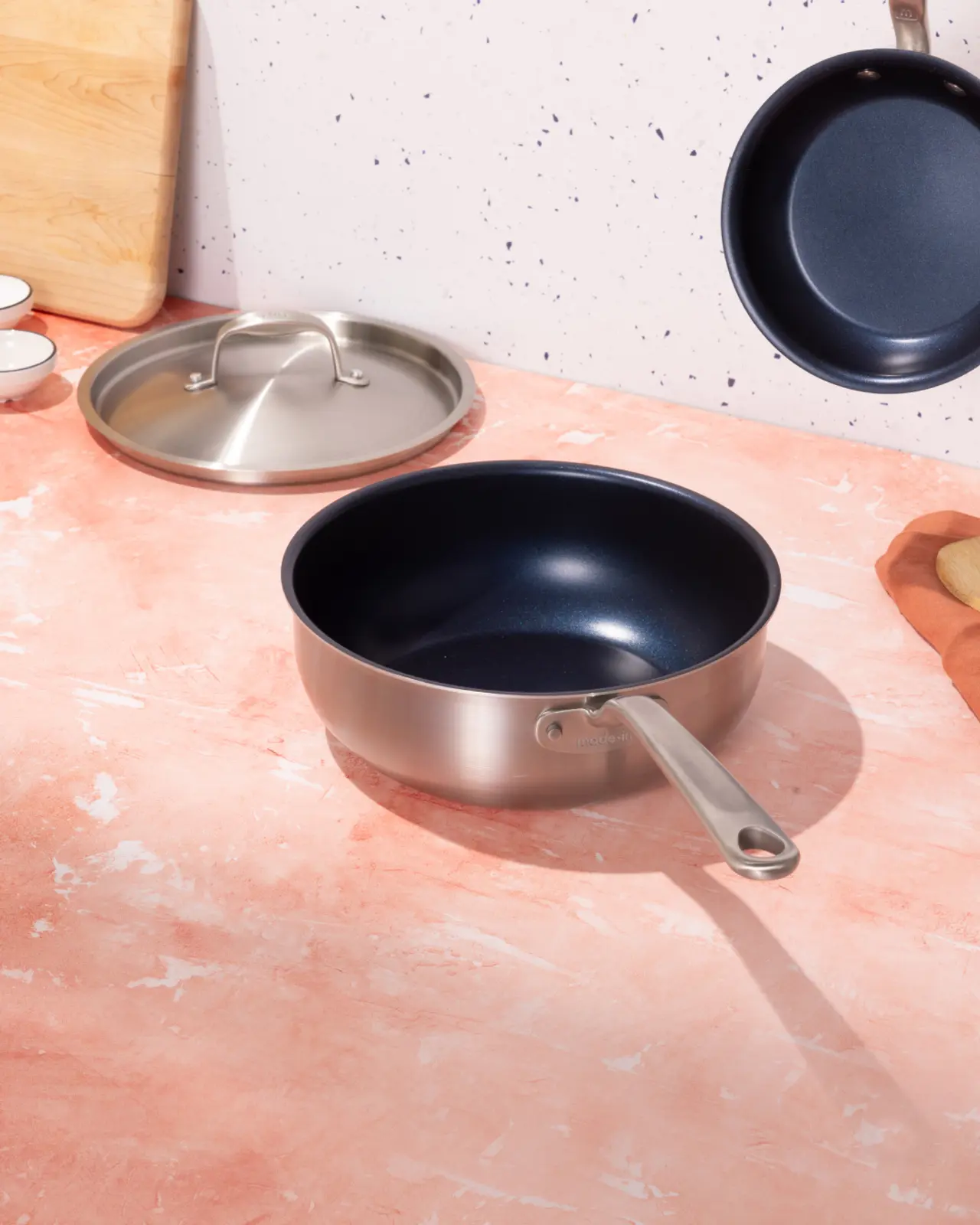 A stainless steel sauté pan with a lid off to the side sits on a terracotta-hued countertop, accompanied by a flipped frying pan and cooking utensils in the background.