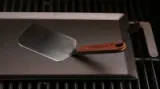 A metal spatula with a wooden handle rests on a dark non-stick baking tray over a grill.