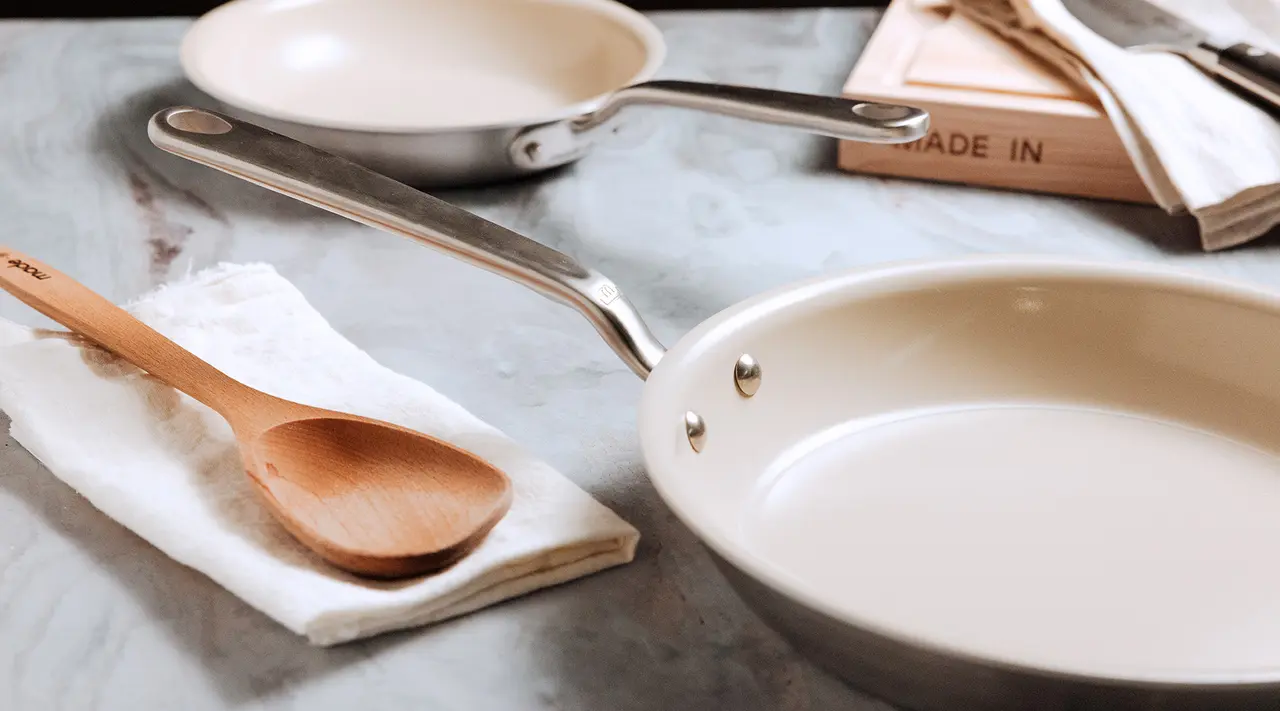 Two empty ceramic pans sit on a countertop beside a wooden spoon resting on a white cloth.