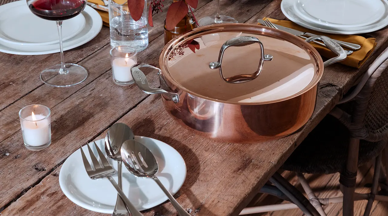 A copper pot on a rustic wooden table set with plates, cutlery, wine glasses, and candles for a cozy meal.