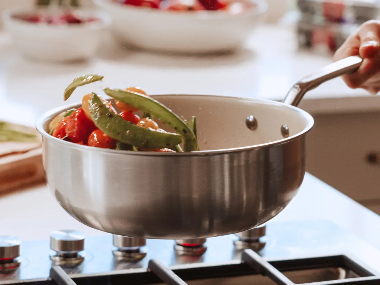 A hand holds a stainless steel pan containing colorful vegetables, being lifted above a stove in a bright kitchen.