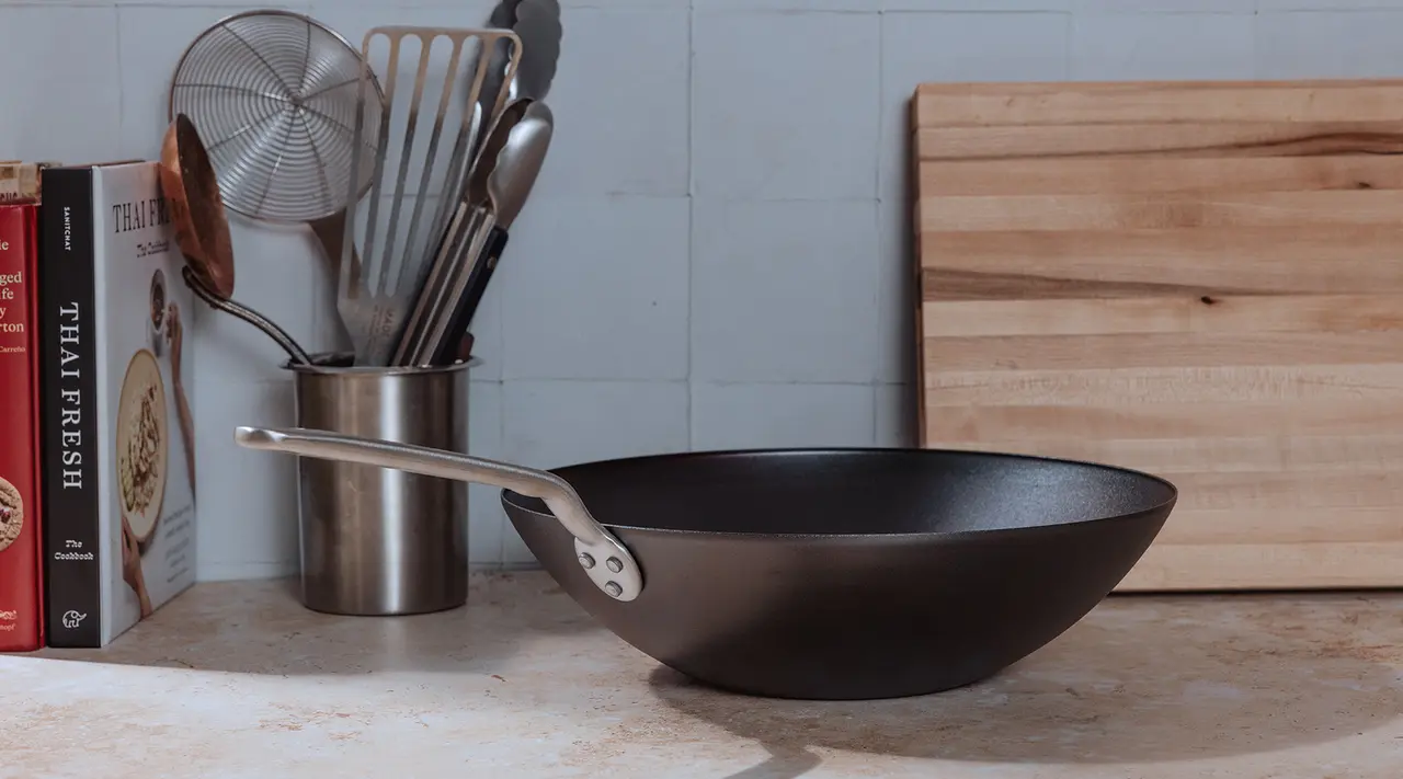 A black cooking pan is placed on a countertop beside a wooden cutting board and various kitchen utensils.