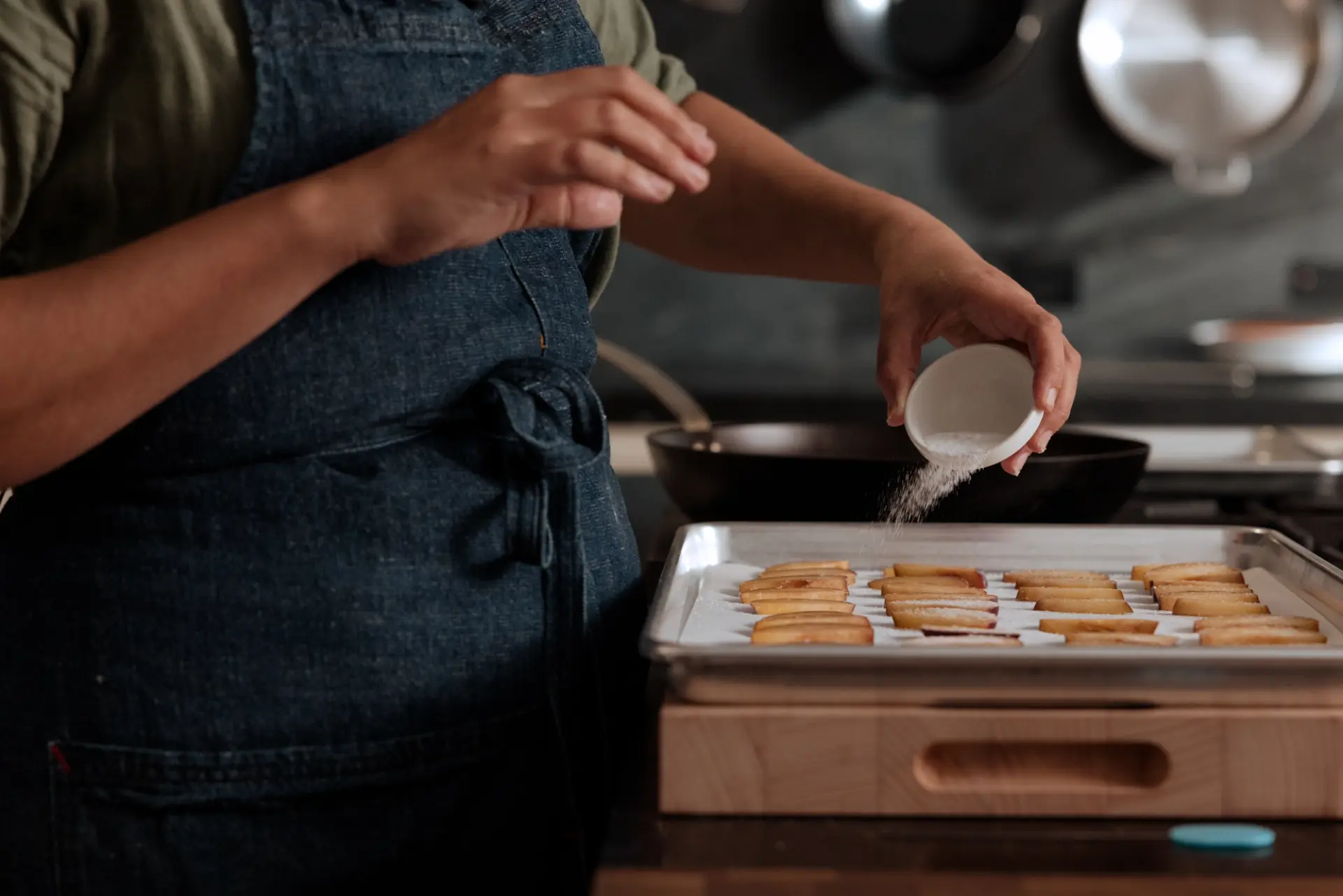 A person wearing an apron is sprinkling sesame seeds over dough on a baking tray in a kitchen setting.